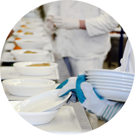 Foto von Mitarbeitenden in weisser Arbeitskleidung und Handschuhen, die in einer Grossküche weisse Teller mit Speisen befüllen und stapeln.