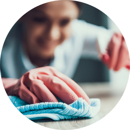 Nahaufnahme einer Person mit rosafarbenen Gummihandschuhen, die mit einem blauen Mikrofasertuch eine Oberfläche reinigt, im Hintergrund verschwommen ein lächelndes Gesicht.